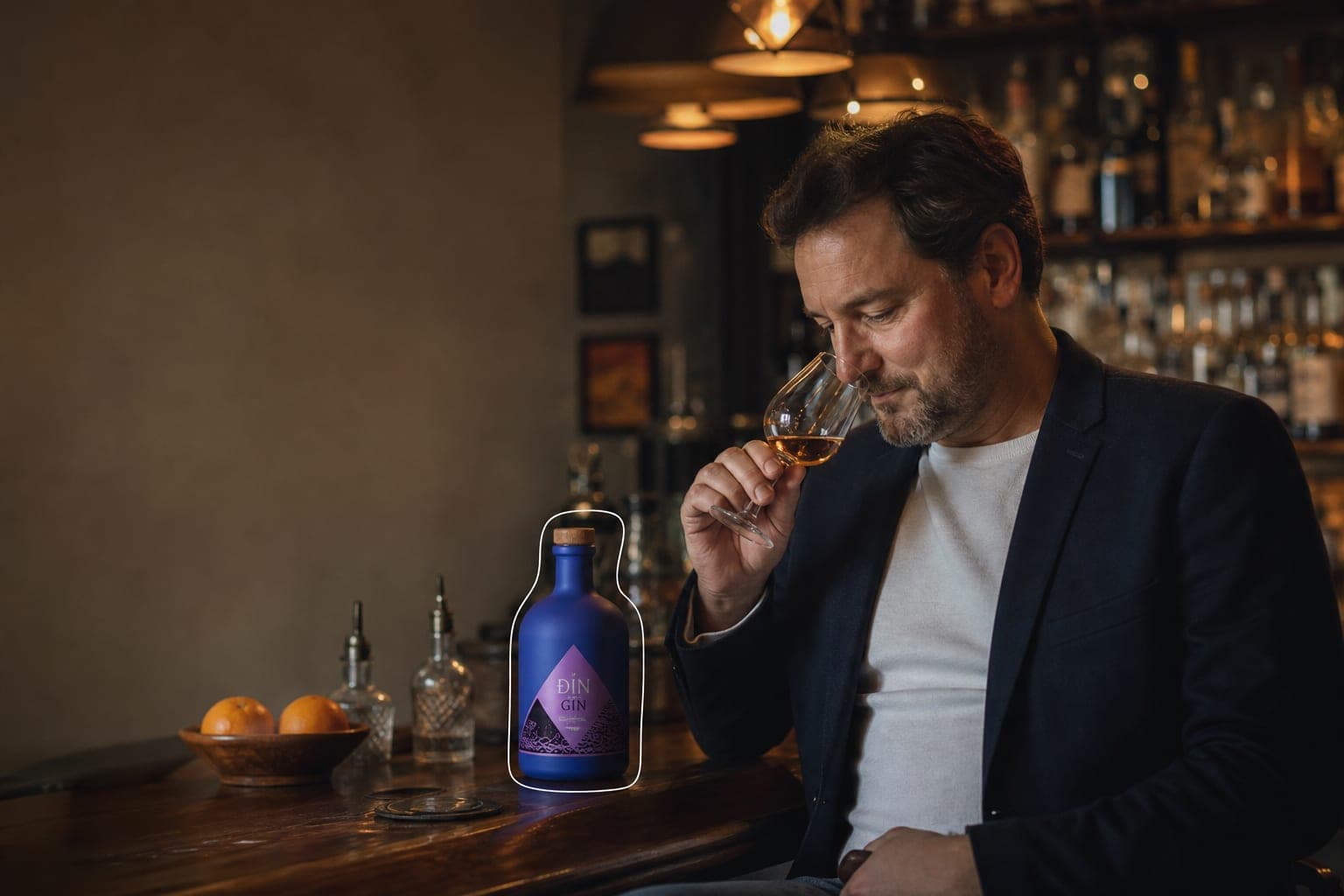 A man at a bar nosing a glass of spirits, a bottle on the counter beside him.